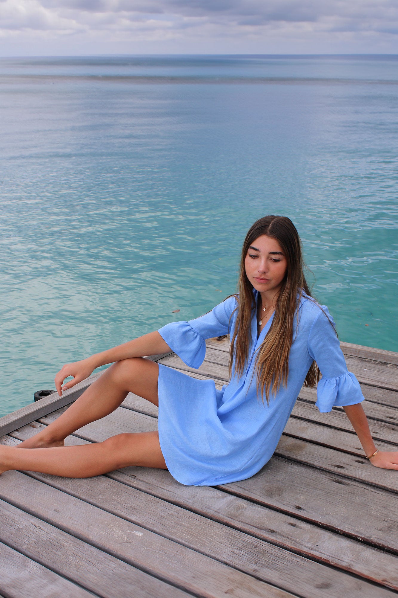 Sitting on the dock in womens pure linen Decima dress with gathered sleeves in plain French blue